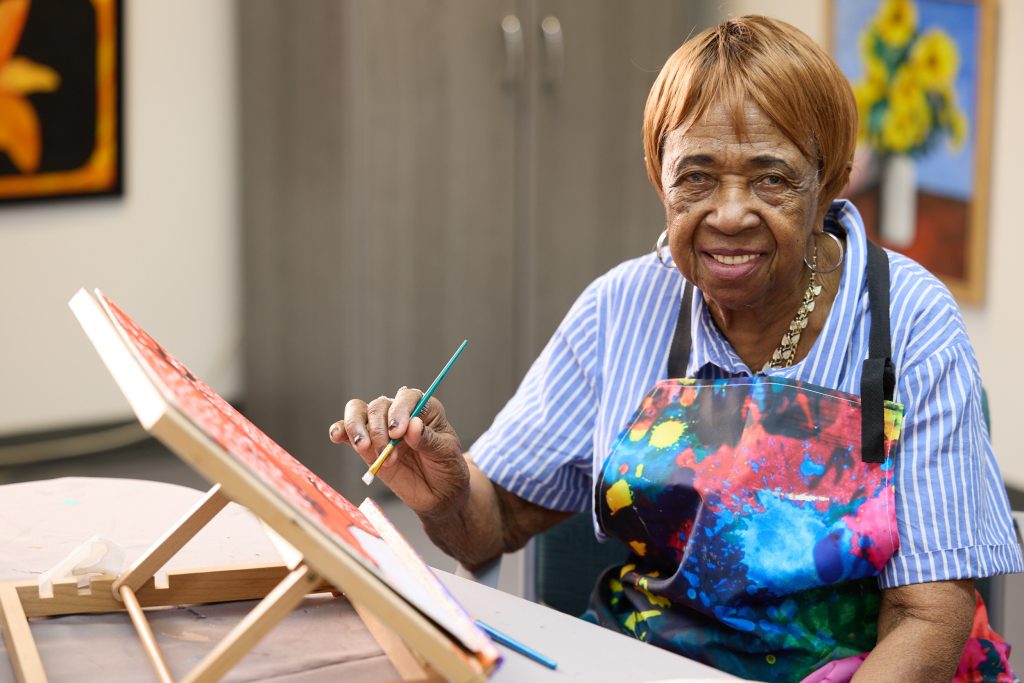 Senior resident painting on an easel in a bright room.