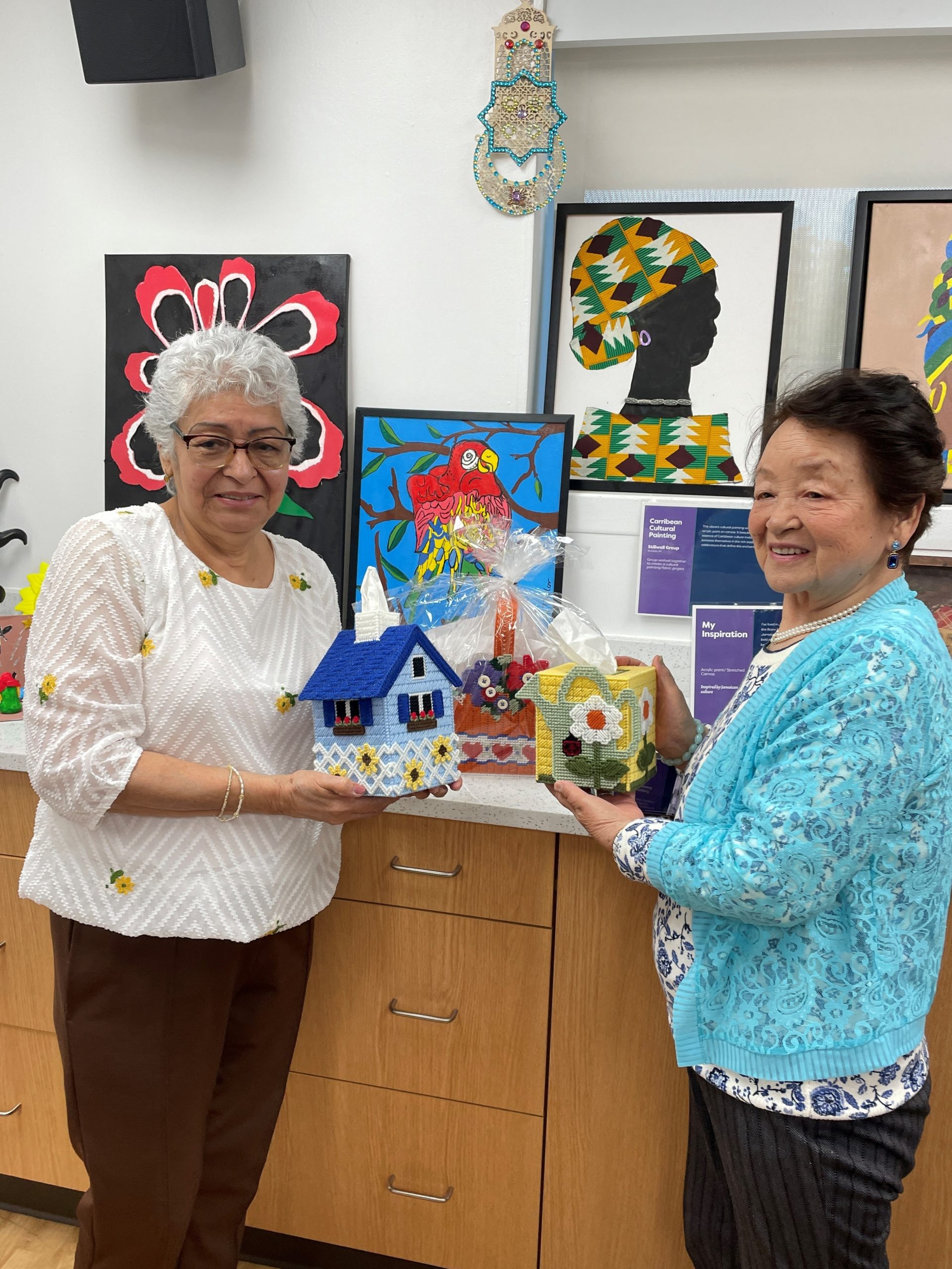 Two women holding colorful knitted houses in a common area.