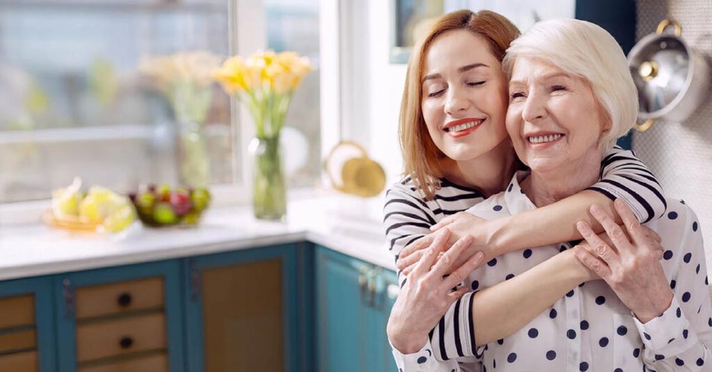 A caregiver hugging a smiling senior woman in a bright kitchen