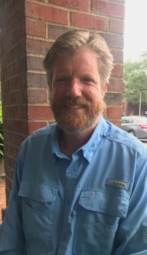 Smiling man outdoors near a brick wall.