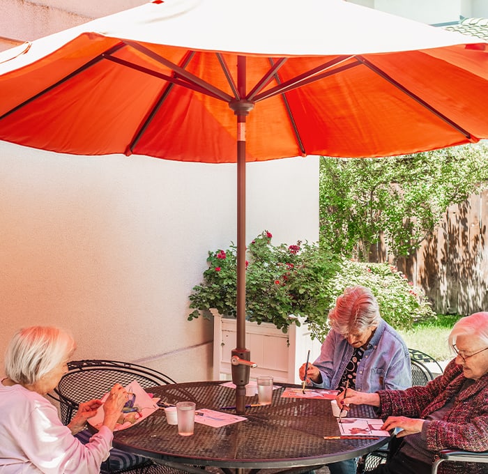Seniors engaging in art activities under a patio umbrella.