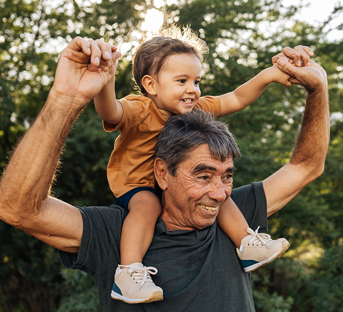 A senior man joyfully carrying a child outdoors.