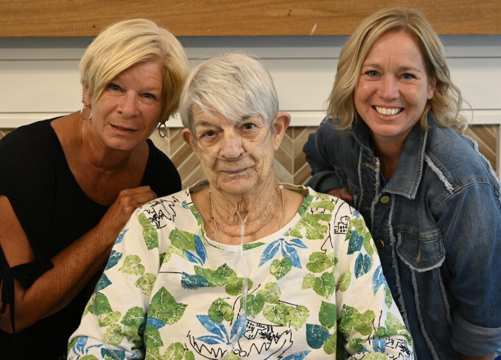Three individuals smiling together, one woman in a floral top with oxygen.
